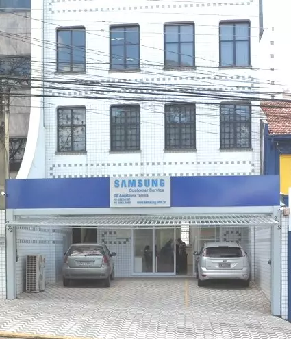 Foto da fachada da Lasan com a placa da Samsung Customer Service depois da ampliação e reforma. A entrada do prédio possui uma cobertura azul e há dois carros estacionados à frente.