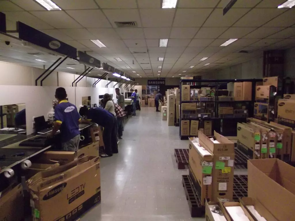Estoque com várias caixas e produtos eletrônicos que estão organizados ao longo do corredor. Há funcionários vestindo camisetas azuis trabalhando com os itens.
