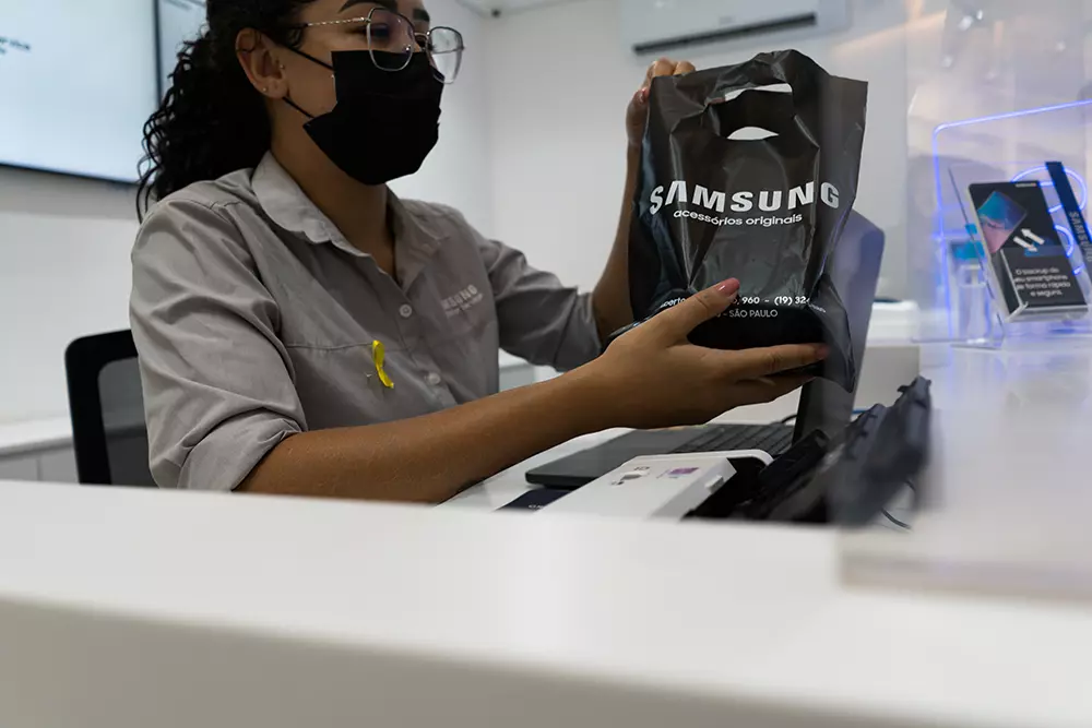 Funcionária da Samsung atrás de um balcão de atendimento. Ela está vestindo uma camisa cinza com o logotipo da empresa e uma máscara facial preta. A funcionária segura uma sacola preta com o logotipo da Samsung.
