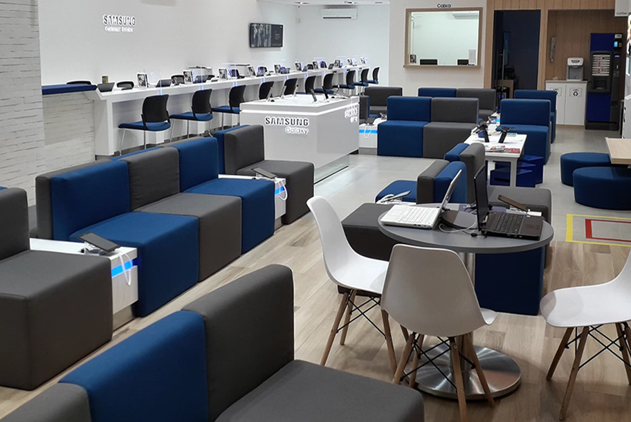 Foto interna da área de atendimento Lasan com vários sofás nas cores azul e cinza e cadeiras em branco. Há balcões de atendimento com computadores e materiais promocionais da Samsung.