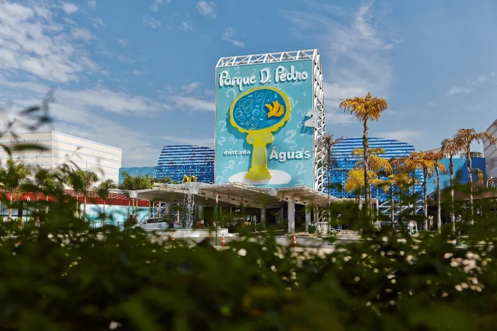 A imagem mostra a entrada do Shopping Parque D. Pedro Shopping. Na frente há uma área paisagística com vegetação, flores brancas em primeiro plano e árvores ao fundo.